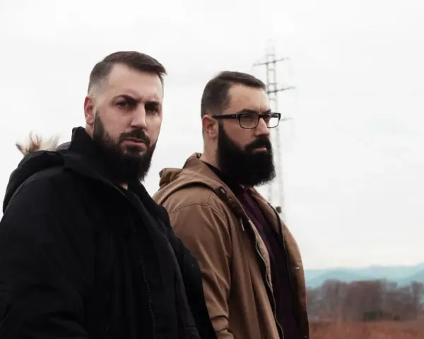 Dos hombres con barba posan con seriedad. Uno viste chaqueta negra, el otro marrón. Parecen listos para un rap de "falsa alarma".