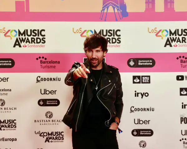 Hombre con chaqueta de cuero negro señala a la cámara en una alfombra roja de Los40 Music Awards. ¡Parece que ha pescado el premio!