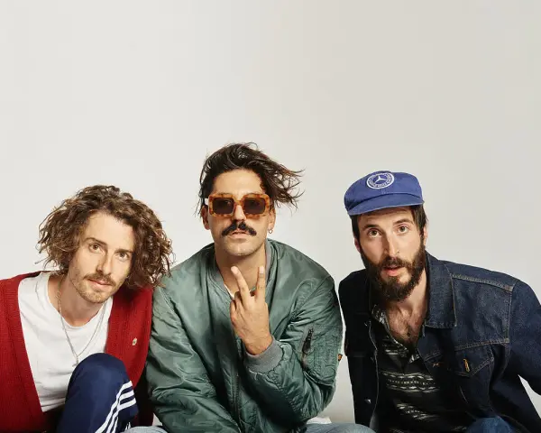 Tres hombres posan, uno con cárdigan rojo, otro con gafas de sol y chaqueta bomber, y el tercero con gorra azul. Parecen un grupo de música, quizás el cantante sexy de Zebras.
