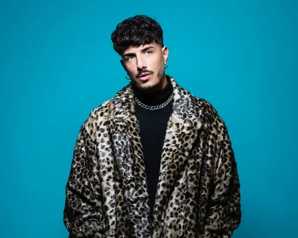 Wade DJ, con su abrigo de leopardo y cadena plateada, posa con confianza ante un fondo azul vibrante.
