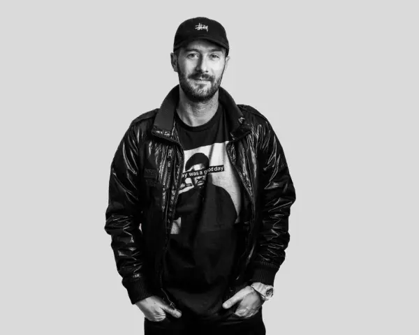 DJ Plan B, con gorra y chaqueta de cuero, sonríe con una camiseta que dice "Today was a good day".