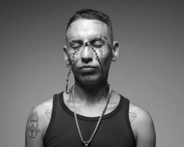 Un joven con tatuajes en la cara y el cuerpo, luciendo un collar de cuentas y una cadena de diamantes. Su estilo neo pistea barcelona es inconfundible.