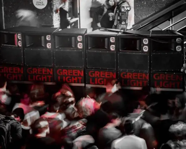 Altavoces apilados con luces rojas "GREEN LIGHT" y gente borrosa bailando. Un sistema de sonido que parece dar problemas.