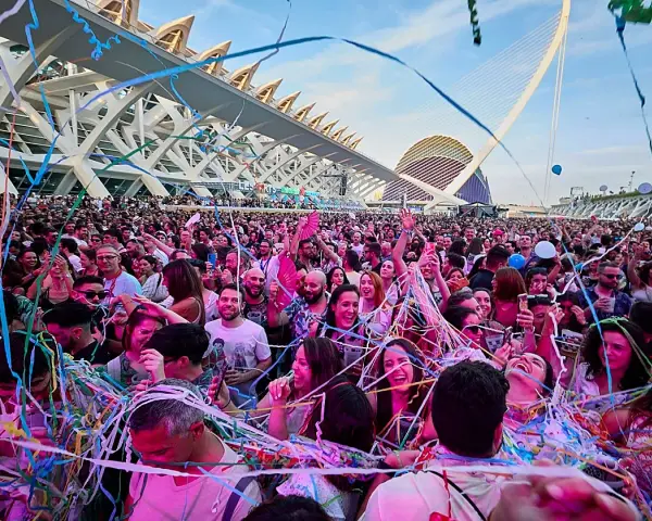 Multitud disfrutando de un fan music fest bajo confeti y luces, con arquitectura moderna de fondo.