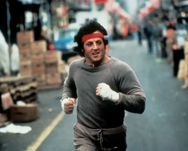 Rocky Balboa corre por la calle, con su icónica banda roja en la cabeza. La banda sonora de Rocky resuena en tu mente.