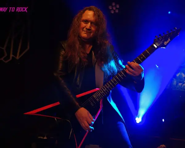 Guitarrista con pelo largo y chaqueta de cuero, tocando una guitarra eléctrica negra con detalles rojos. El escenario está iluminado con luces azules, creando un ambiente de rock.