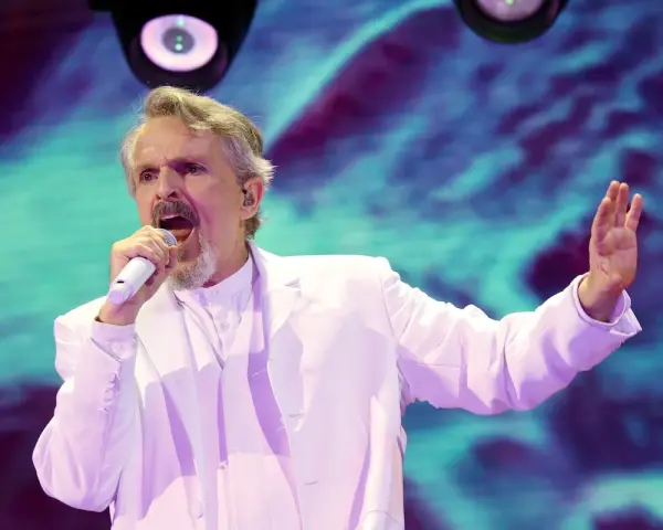 Un hombre con traje blanco canta en un escenario con luces vibrantes. ¡Murcia on!