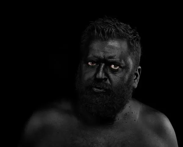 Hombre con el rostro y cuerpo cubiertos de pintura negra, ojos penetrantes. Parece un guerrero ancestral, don fluor.