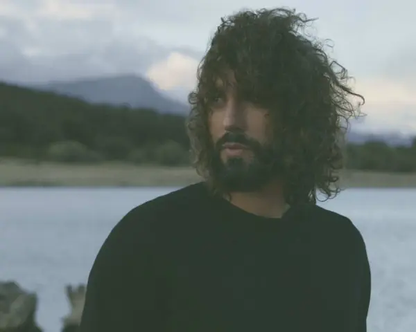 Un hombre con cabello rizado y barba mira hacia el horizonte, entre el miedo y el paraíso de un paisaje acuático y montañoso.
