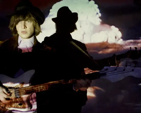 Jacco Gardner, con sombrero y chaqueta, toca el bajo frente a una proyección psicodélica.