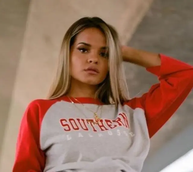 Aleesha Rose, con su cabello rubio y suéter gris con mangas rojas, posa con confianza.