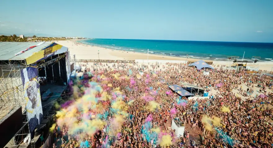 Multitud vibrante en el Arenal Sound Valencia, lanzando polvo de colores bajo el sol. El mar azul y la playa de fondo crean un escenario festivo.