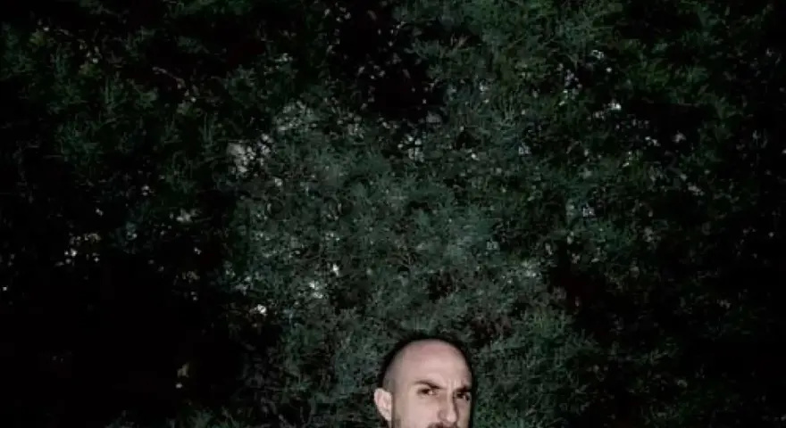 Un hombre calvo, con mirada intensa, posa frente a un fondo de follaje oscuro. Podría ser un sicario rapero, listo para soltar rimas letales.