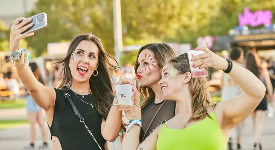 Tres amigas con maquillaje festivo y bebidas en un festival de verano.