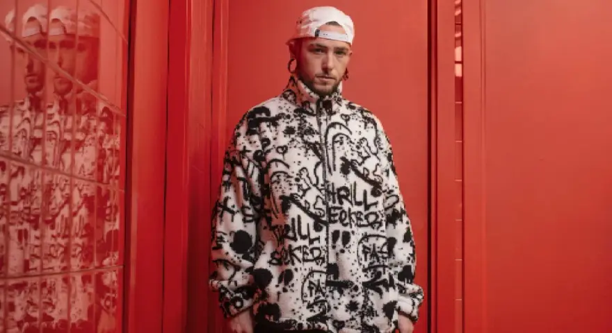 El cantante Kaze, con gorra blanca y chaqueta polar con grafitis, posa en un pasillo rojo vibrante.