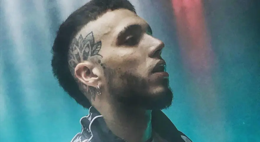 Un joven **hard cantante** con tatuaje en la cabeza y barba, mirando hacia arriba con luces de neón de fondo.