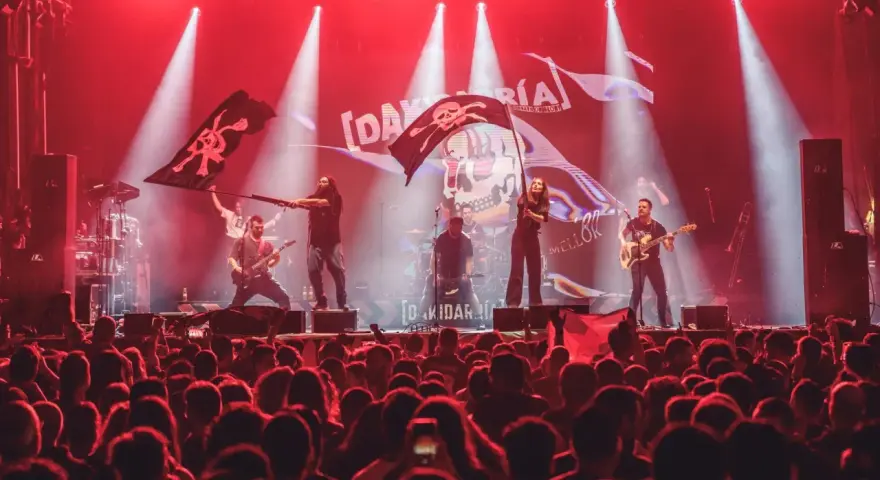 Una banda toca en un escenario iluminado con luces rojas, ondeando banderas piratas. El público, con trece roeis, anima la actuación.