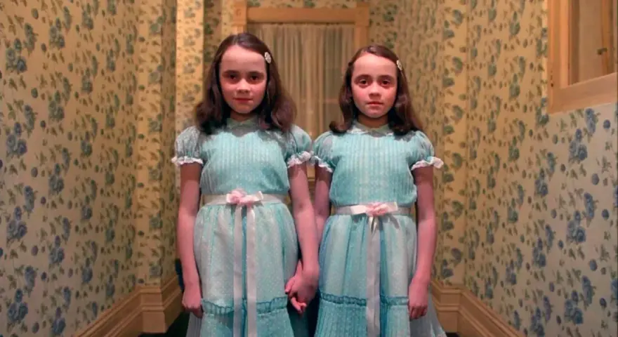 Dos niñas idénticas, vestidas con trajes azules, se toman de la mano en un pasillo con papel tapiz floral. Son las gemelas de "Bso El Resplandor".