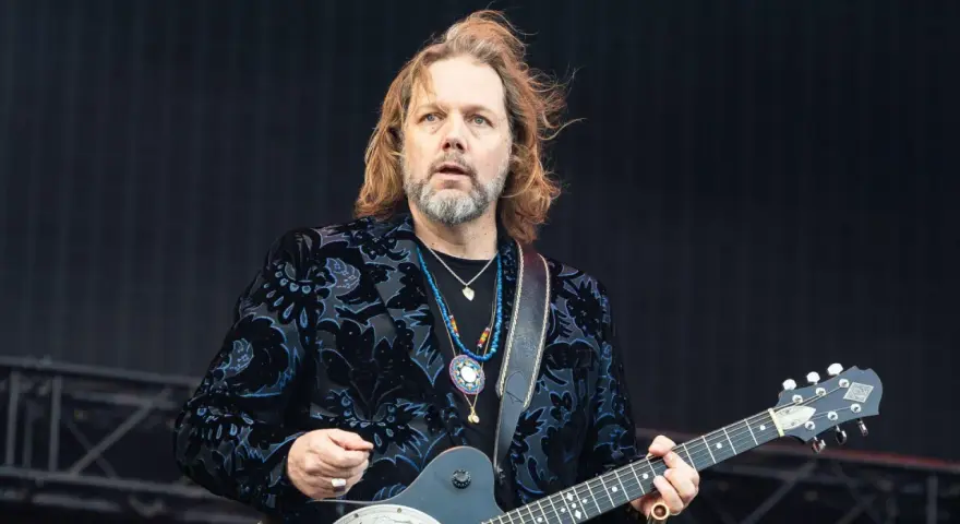 Rich Robinson, guitarrista con cabello largo y barba, luce un saco negro con estampado floral azul y varias cadenas.