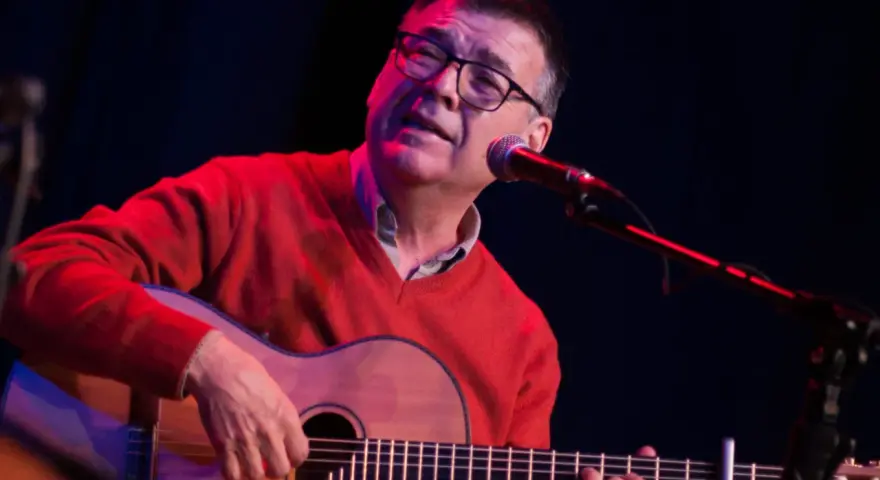 Mario Díaz, con gafas y un suéter rojo, toca la guitarra y canta frente a un micrófono en un escenario con luces tenues.