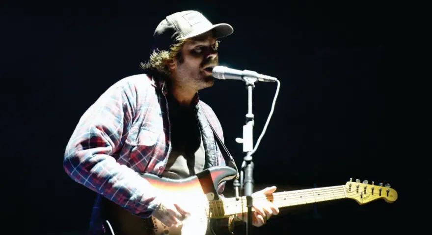 Neil Halstead, con gorra y camisa a cuadros, toca la guitarra y canta en un concierto.