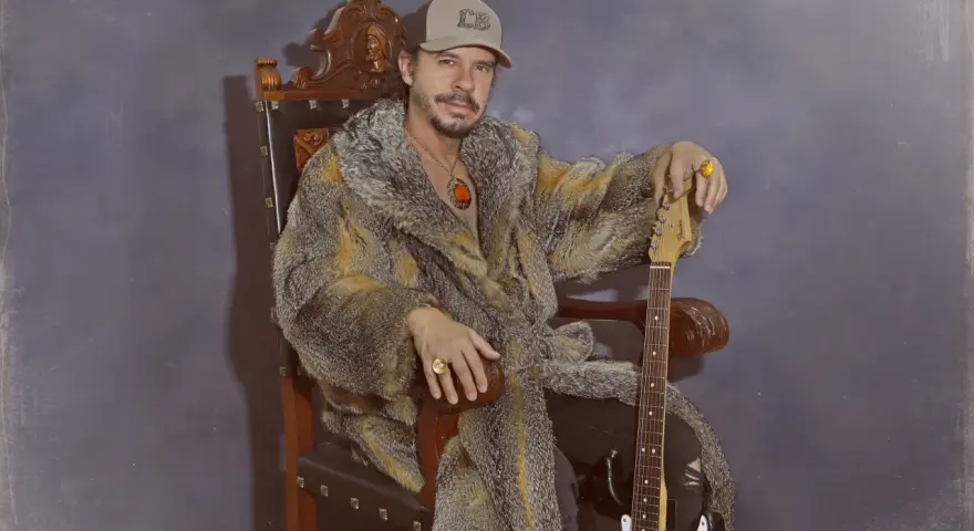 Luis Brea, con gorra y abrigo de piel, sentado en un trono con una guitarra.