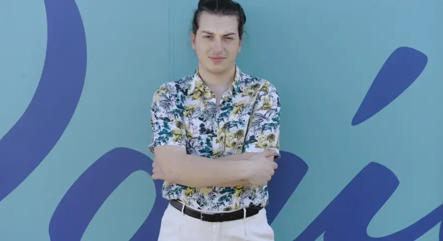 Joven con camisa tropical y pantalones claros, brazos cruzados, frente a un mural azul con formas orgánicas. Parece un miembro del ortiga grupo.