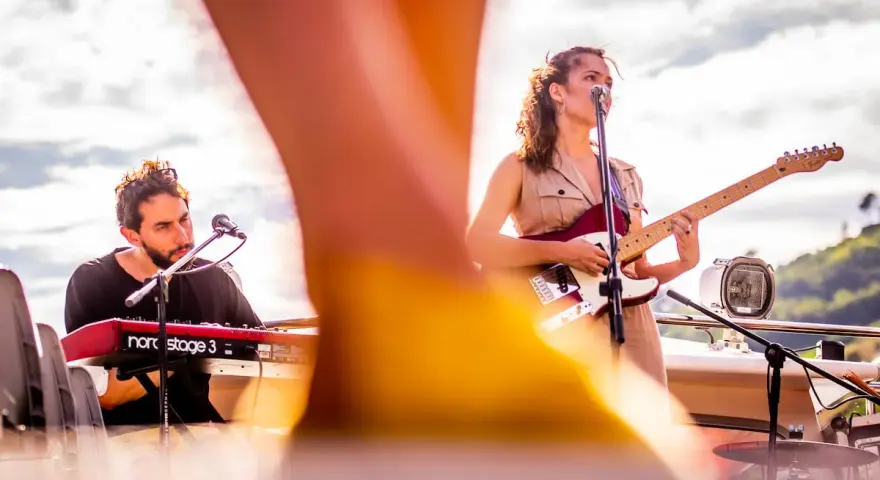 Maria y Feu, una banda en vivo, tocan en un escenario al aire libre. Ella canta y toca la guitarra, él en los teclados.
