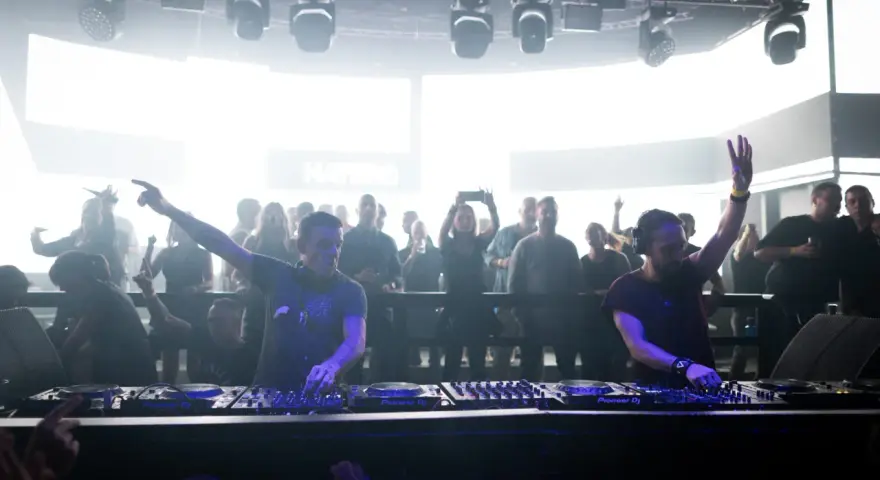 Dos DJs actuando en un club con público al fondo.