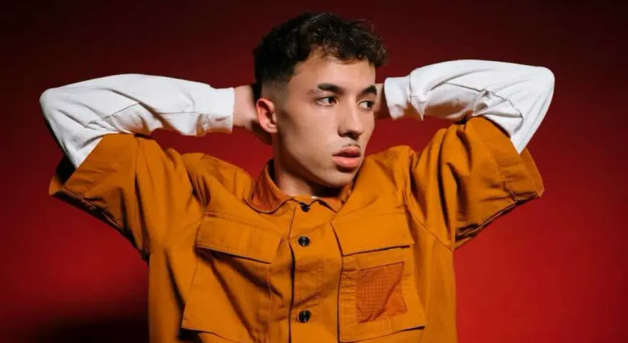 Joven con las manos detrás de la cabeza, vistiendo una chaqueta naranja y una camiseta blanca. Parece un pole cantante listo para actuar.