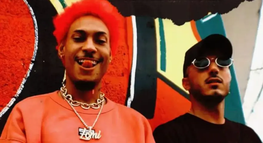 Dj Ballesteros y su colega posan frente a un mural vibrante. El primero, con cabello rojo y un collar dorado, sonríe con la lengua afuera.