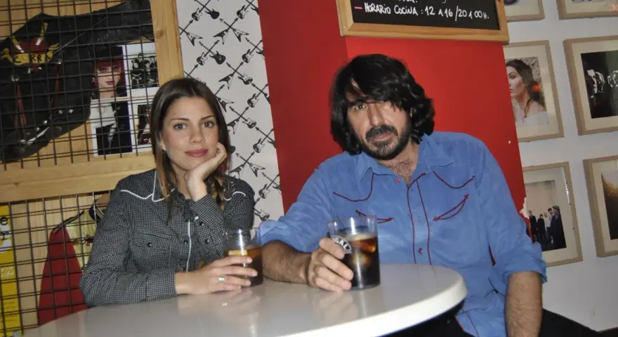 Pareja en un bar con decoración rockera. Ella, con camisa de cuadros, apoya la barbilla en la mano. Él, con camisa azul, sostiene un vaso. La furia trinidad de la música inspira el ambiente.