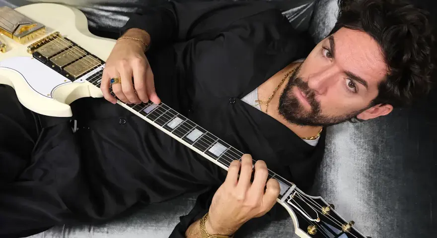 El cantante Siloe, con barba y cabello oscuro, toca una guitarra eléctrica blanca con detalles dorados, recostado en un sofá de terciopelo gris.