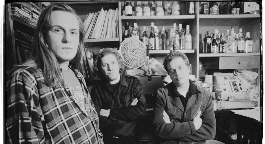 Tres hombres posan en blanco y negro, con estanterías de discos y botellas detrás.