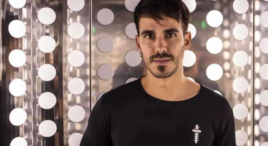 Raúl Pacheco, DJ, posa con una camiseta negra frente a un fondo metálico con círculos blancos brillantes.