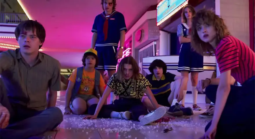 Los personajes de Stranger Things, con Eleven al frente, sentados en el suelo de un centro comercial, evocando las icónicas canciones de la serie.