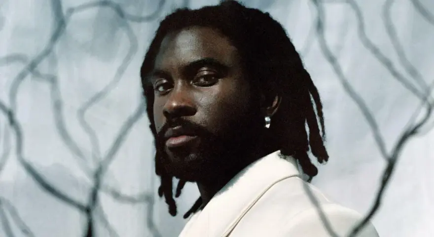 Retrato de un rapero español con rastas y un pendiente plateado, vistiendo una chaqueta clara.