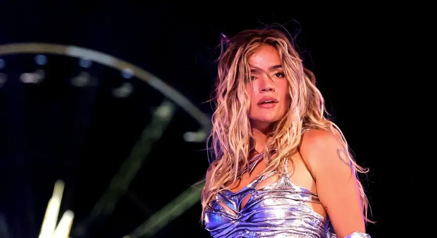 Karol G, con su melena rubia al viento, brilla en el escenario. Su atuendo plateado y el ambiente nocturno sugieren un concierto inolvidable de su gira.