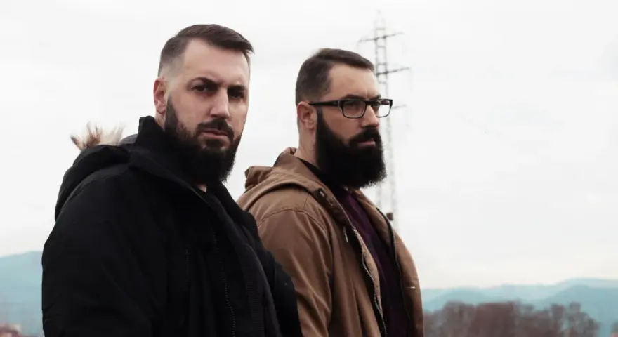 Dos hombres con barba posan con seriedad. Uno viste chaqueta negra, el otro marrón. Parecen listos para un rap de "falsa alarma".