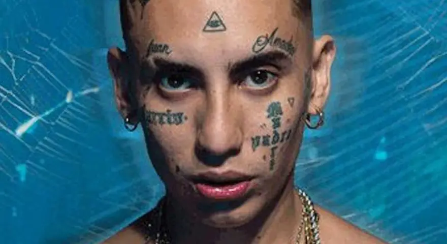 Joven con tatuajes en la cara, incluyendo un ojo dentro de un triángulo, y cadenas de oro. Su estilo neo pistea madrid es inconfundible.