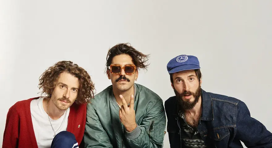 Tres hombres posan, uno con cárdigan rojo, otro con gafas de sol y chaqueta bomber, y el tercero con gorra azul. Parecen un grupo de música, quizás el cantante sexy de Zebras.