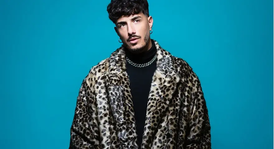 Wade DJ, con su abrigo de leopardo y cadena plateada, posa con confianza ante un fondo azul vibrante.