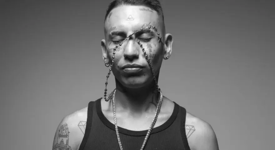 Un joven con tatuajes en la cara y el cuerpo, luciendo un collar de cuentas y una cadena de diamantes. Su estilo neo pistea barcelona es inconfundible.