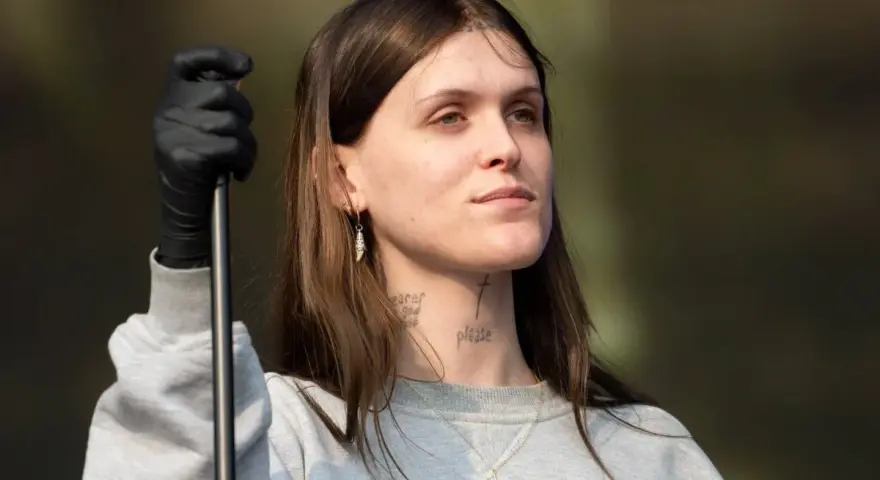 Ethel Cain, con guantes negros y un collar de cruz, mira hacia un lado con una expresión pensativa.