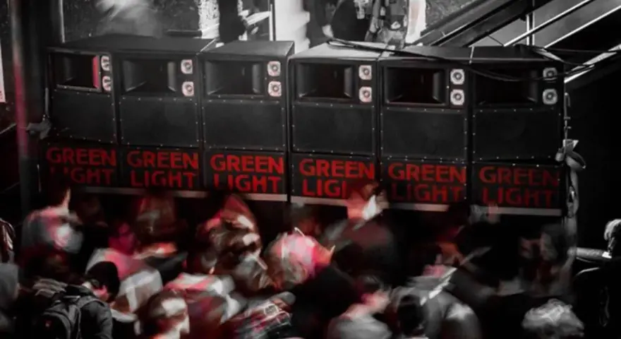 Altavoces apilados con luces rojas "GREEN LIGHT" y gente borrosa bailando. Un sistema de sonido que parece dar problemas.