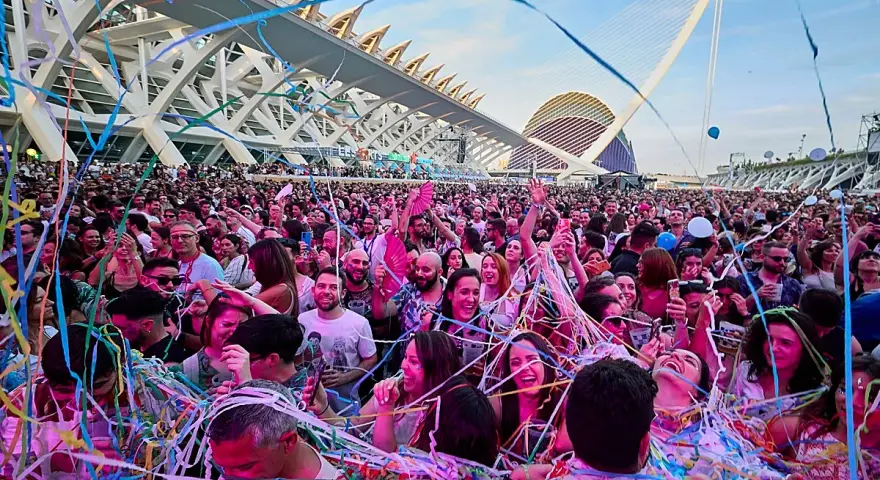 Multitud disfrutando de un fan music fest bajo confeti y luces, con arquitectura moderna de fondo.