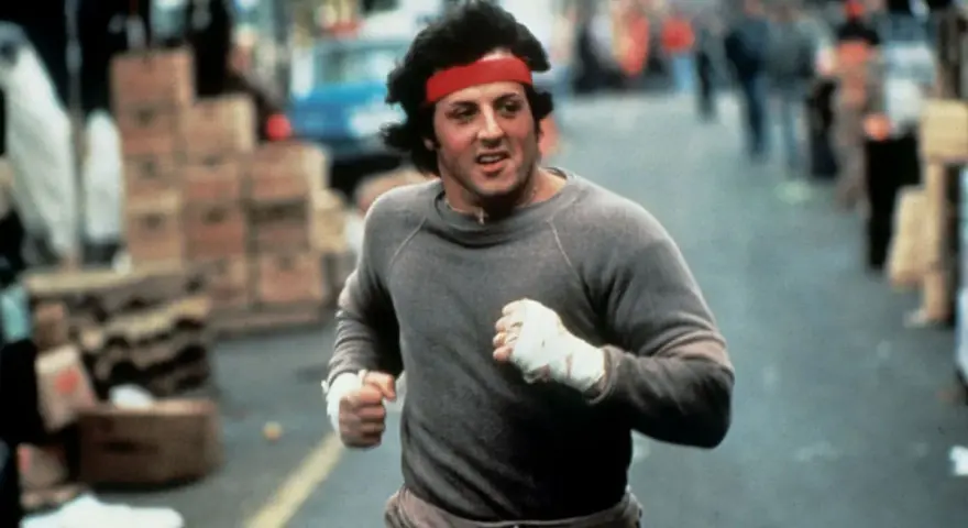 Rocky Balboa corre por la calle, con su icónica banda roja en la cabeza. La banda sonora de Rocky resuena en tu mente.