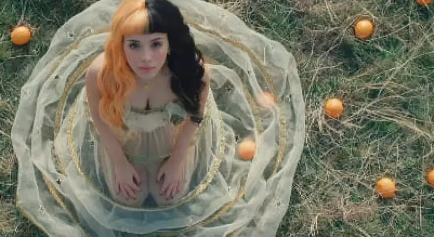 Melanie Martinez, con su icónico cabello mitad naranja y mitad negro, rodeada de naranjas en un campo. ¡Atentos a los próximos eventos de Melanie Martinez!