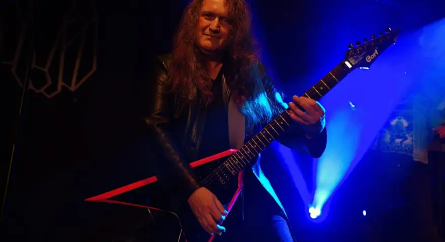 Guitarrista con pelo largo y chaqueta de cuero, tocando una guitarra eléctrica negra con detalles rojos. El escenario está iluminado con luces azules, creando un ambiente de rock.
