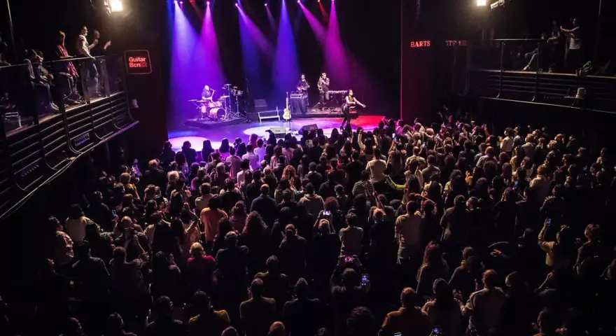 Un concierto en Guitar BCN ilumina el escenario con luces púrpuras mientras una multitud entusiasta graba el momento.